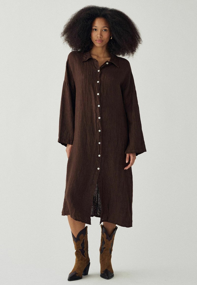 Femme aux cheveux bouclés volumineux portant une longue robe boutonnée marron foncé et des bottes de cowboy marron, debout devant un fond clair uni.