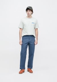 Γκρι βαμβακερό μπλουζάκι με μπλε λογότυπο, μπλε βαμβακερό παντελόνι και καφέ δερμάτινα παπούτσια με κορδόνια. Το μοντέλο στέκεται μπροστά σε λευκό φόντο.