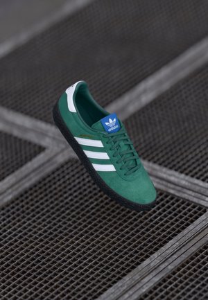 Green suede sneaker with white stripes, featuring a blue tongue logo and a textured black rubber sole. Model name: "Montreal."