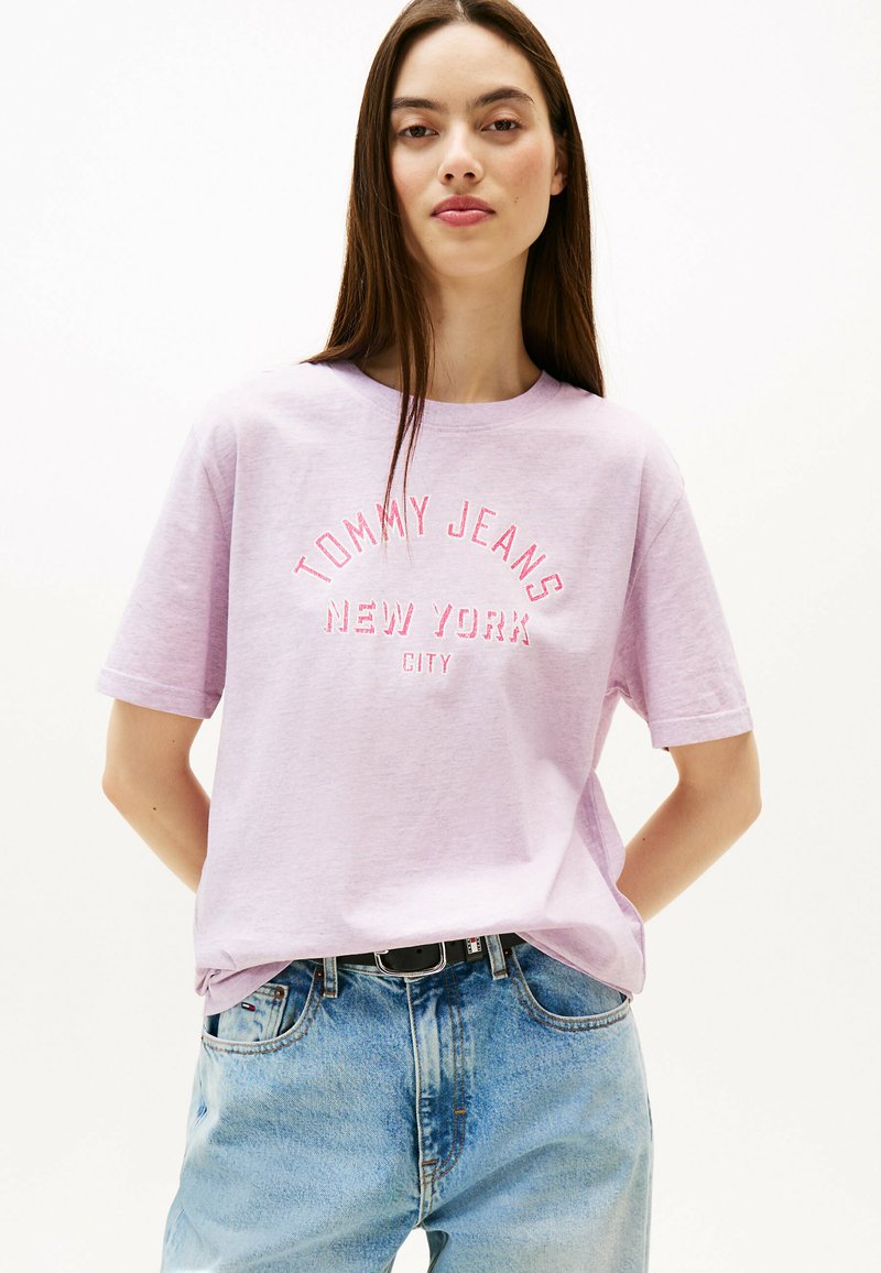 Camiseta de algodón de color púrpura claro con "TOMMY JEANS NEW YORK CITY" en texto bordado rojo. Mangas cortas y cuello redondo. Corte casual.