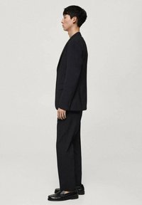 Mørk blazer og matchende bukser lavet af fint tekstureret stof. Skoene har skinnende sorte loafers. Enkel, skræddersyet design.