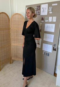 Fekete ruha V-nyakkal, rövid ujjakkal, bugyros oldalakkal és combközépig érő hasítékkal. Textúrált háttér tervezési vázlatokkal és színmintákkal.