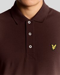 Hnedé polokošeľa z textúrovanej látky, s golierom so štyrmi gombíkmi a malým vyšívaným žltým logom vtáka na ľavej strane hrude.