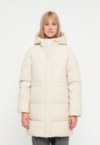 Beige polstret jakke med hætte, der har lynlås lukning og syede sektioner. Designet har en afslappet pasform og lange ærmer.
