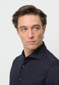 Marineblaues Hemd mit spitzem Kragen und dunklen Knöpfen, mit einer glatten Textur und taillierter Passform, präsentiert an einem Mann mit heller Haut.