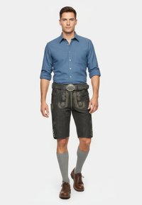 Man som bär en blå skjorta med knappar, svarta lädershorts med broderade detaljer, grå knästrumpor och bruna snörskor.