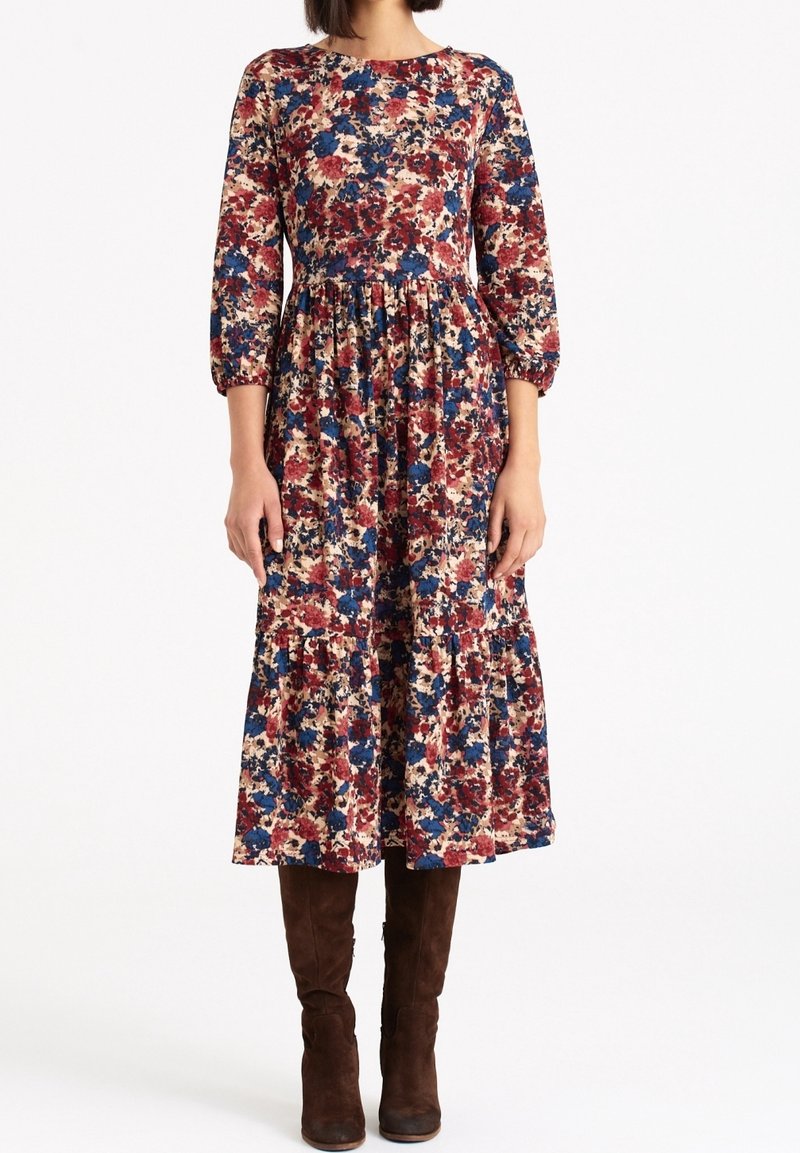 Robe fleurie avec manches longues bouffantes en nuances de rouge, bleu et beige ; taille froncée et jupe à volants. Le mannequin porte des bottes hautes marron au genou.