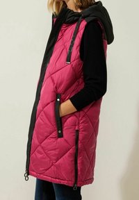 Ärmlös vadderad väst i klar rosa med en svart huva. Har sidofickor med dragkedja, diamantmönster och kontrasterande svart kant.