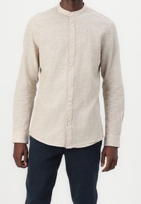 Camisa de manga larga beige con cuello redondo, con botones al frente y tejido texturizado. Se lleva con pantalones oscuros, mostrando un ajuste relajado.