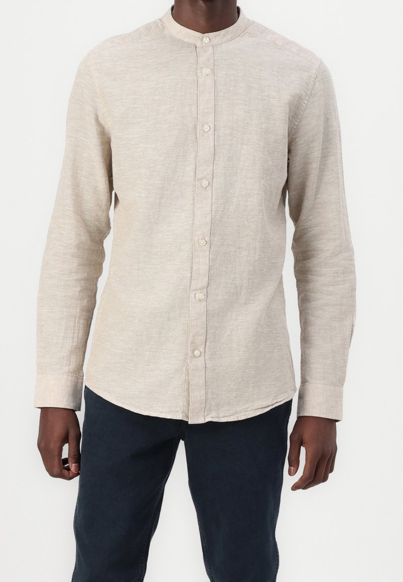 Camisa de manga larga beige con cuello redondo, con botones al frente y tejido texturizado. Se lleva con pantalones oscuros, mostrando un ajuste relajado.