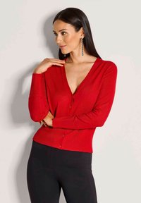 Roter Cardigan mit tiefem V-Ausschnitt, geripptem Material und Knopfverschluss. Kombiniert mit schwarzen, figurbetonten Hosen, die die schlanke Silhouette betonen.
