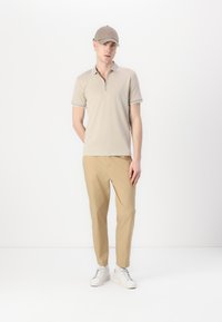 Beige poloskjorte med glidelås og kontrastkrage, sammen med lysebrune bukser og hvite sneakers, satt mot en enkel bakgrunn.