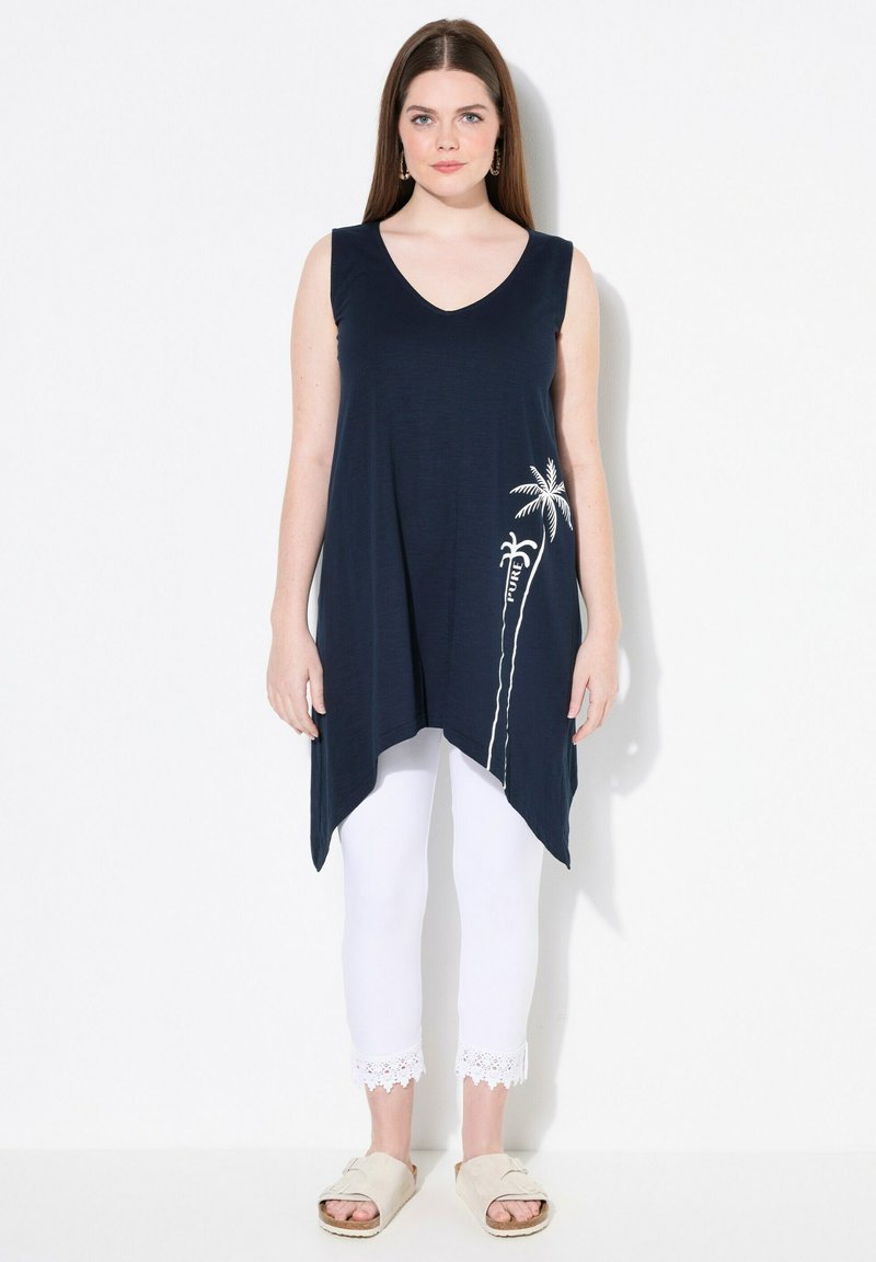 Túnica sin mangas de color marino con escote en V, que presenta un gráfico de palmera en blanco. Combinada con leggings cortos blancos con encaje y sandalias beige.