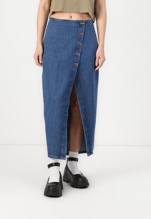 Denim maxi-rok met hoge taille, een split aan de voorkant, houten knoopdetails en een ruwe zoom. In combinatie met zwarte platform schoenen en witte sokken.