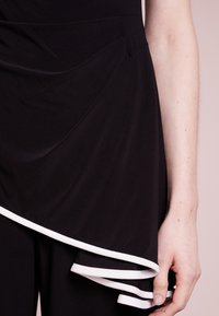 Svart topp med en draperad design, med vit kant längs fållen. Slät textur med lager av tyg. Närbild på en hand som håller tyget.