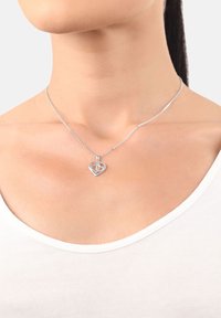 Collana con pendente a forma di cuore in argento con piccole pietre incastonate, indossata al collo di una donna sopra una maglia bianca, su uno sfondo semplice.