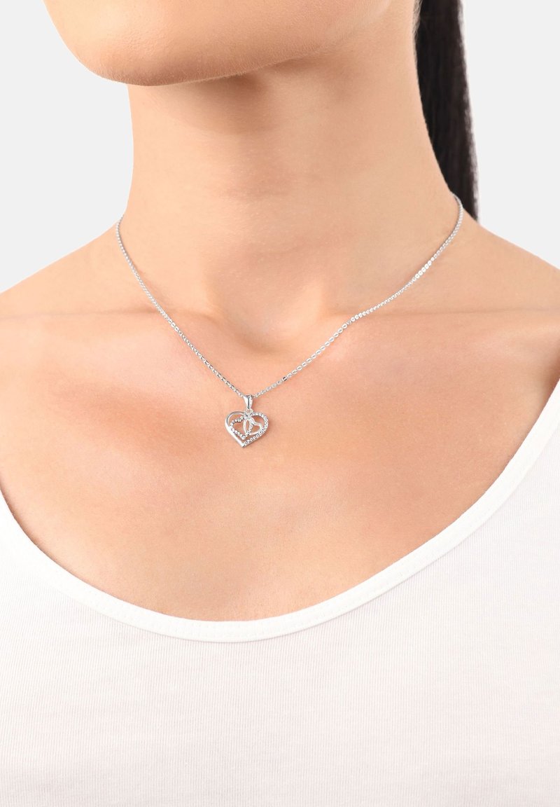 Collana con pendente a forma di cuore in argento con piccole pietre incastonate, indossata al collo di una donna sopra una maglia bianca, su uno sfondo semplice.