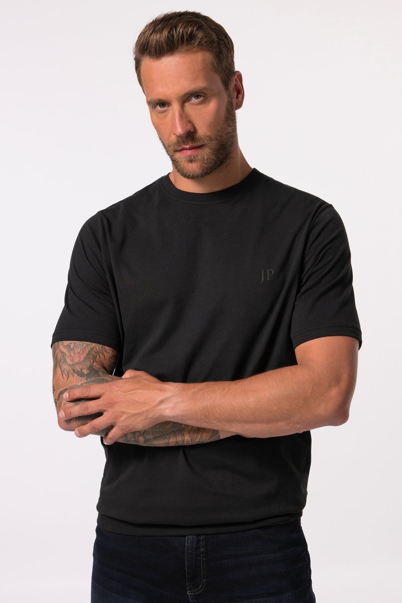 Mann mit Bart, der ein schwarzes Kurzarmshirt trägt, verschränkte tätowierte Arme und schaut direkt nach vorne vor weißem Hintergrund.