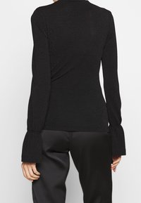 Haut noir à manches longues en tissu texturé scintillant. Présente des poignets évasés et une silhouette ajustée. Associé à un pantalon ample et foncé.