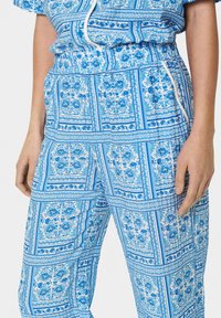 Mėlynos raštuotos kelnės su gėlių raštu ant balto fono, elastinga liemenėle ir šoninėmis kišenėmis. Medžiaga atrodo lengva ir pralaidi orui.