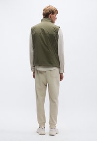 Oliwkowa bezrękawnikowa kamizelka z wysokim kołnierzem, w zestawieniu z kremowymi spodniami. Lekki materiał, luźny krój, prosty design, bez widocznych elementów metalowych.