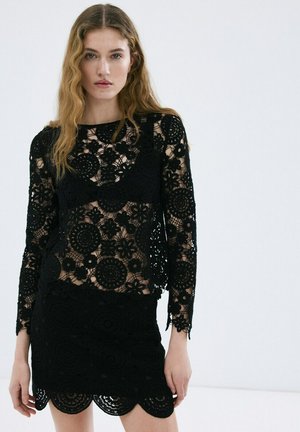 Mujer con cabello ondulado que lleva una blusa de encaje floral negra de manga larga y una falda de encaje negra a juego con dobladillo festoneado.