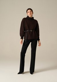 Donna in piedi che indossa un maglione scuro a trama con una cintura nera, pantaloni neri dritti e scarpe nere a punta, contro uno sfondo semplice.