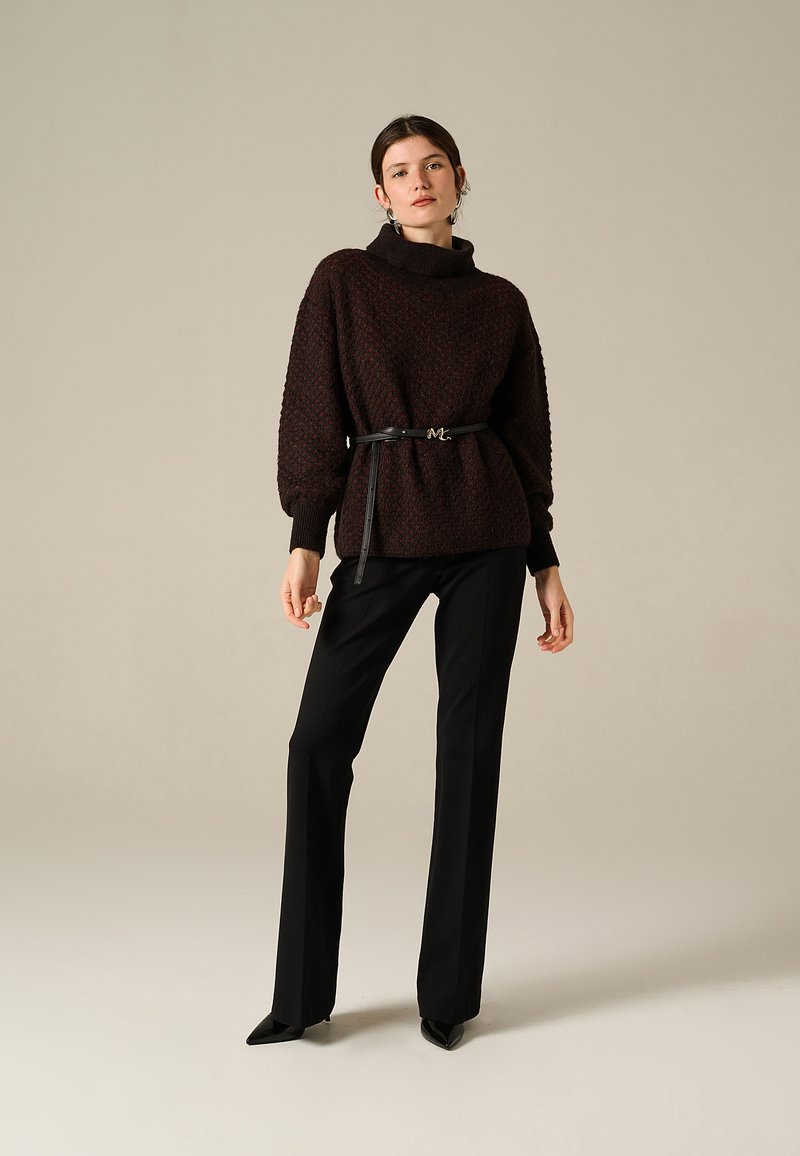 Donna in piedi che indossa un maglione scuro a trama con una cintura nera, pantaloni neri dritti e scarpe nere a punta, contro uno sfondo semplice.