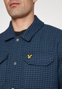 Giacca houndstooth blu navy con colletto bottonato, due tasche frontali e logo giallo ricamato sul petto. Tessuto texturizzato.