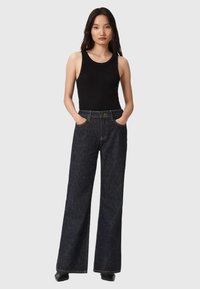 Donna che indossa un top senza maniche nero infilato in un paio di jeans a pieghe a vita alta di colore scuro con un motivo a macchie, con le mani nelle tasche su uno sfondo grigio.