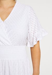 Robe blanche à œillets avec col en V, manches courtes à volants et taille froncée, portée par une personne avec bras et cou visibles.