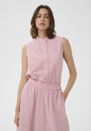 Naine, kellel on pruunid õlgadeni juuksed, seljas varrukateta roosa-valge vertikaalsete triipudega nööpidega kleit, mis on taljast kokku tõmmatud, seisab ühe käega taskus.