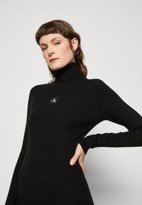 Abito a collo alto nero a coste con maniche lunghe, caratterizzato da una patch con logo sul petto e una vestibilità slim, realizzato in tessuto texturizzato.