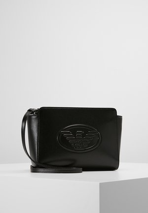Petit sac bracelet en cuir noir avec logo rond embossé à l'avant, forme rectangulaire et fine bandoulière, posé sur une surface blanche avec un fond clair.