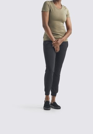 Kobieta stojąca ze splecionymi rękami, ubrana w dopasowany khaki t-shirt, czarne spodnie dresowe i czarne buty wsuwane, na jednolitym tle.