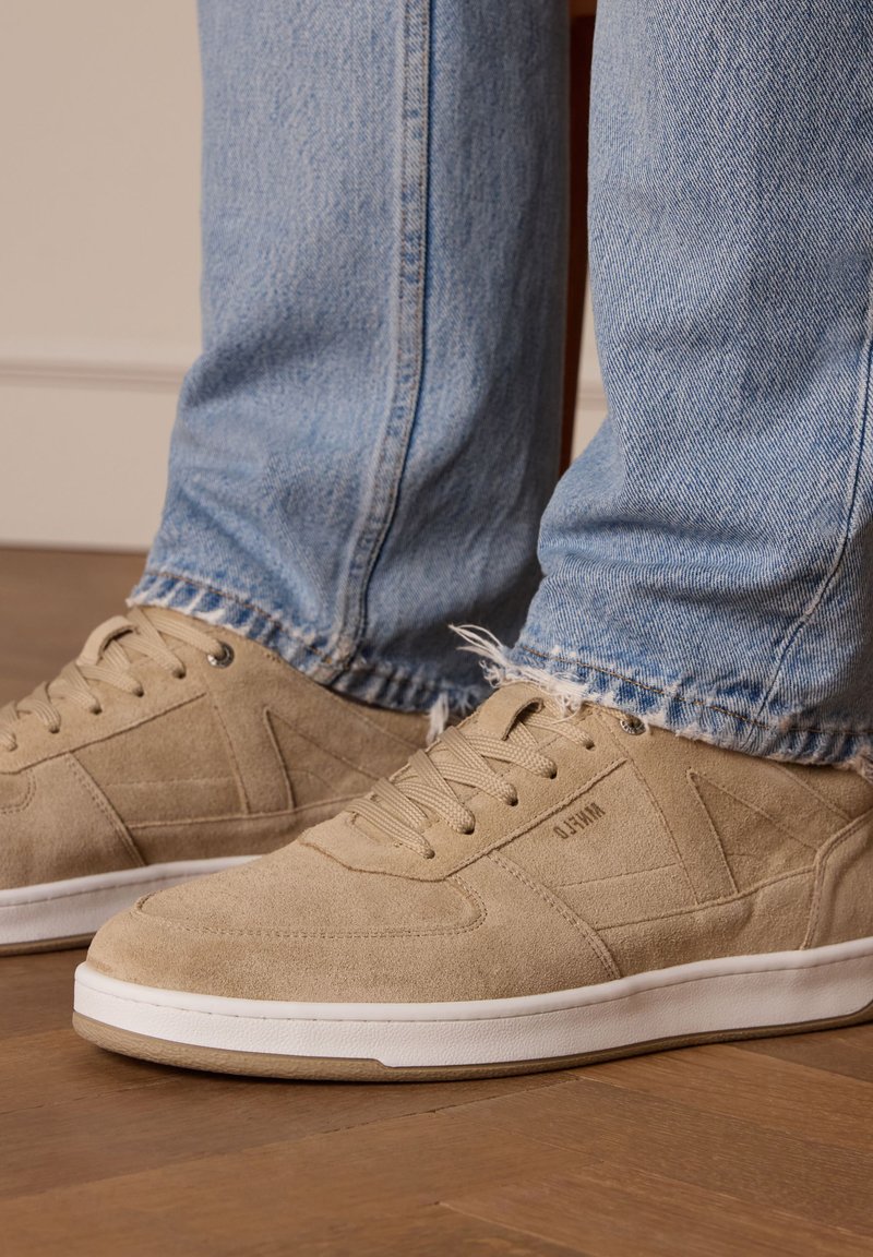 Sneaker beige in suede con una texture liscia, suola in gomma bianca e dettagli di design geometrico sottili. Indossati con jeans in denim blu sfilacciati.