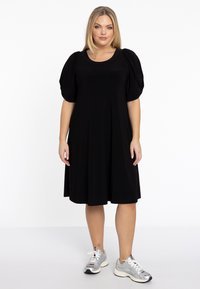 Schwarzes kurzärmeliges Kleid aus weichem Stoff, mit Rundhalsausschnitt, gerafften Puffärmeln und einem schwingenden Rock. Kombiniert mit silbernen Sneakers.