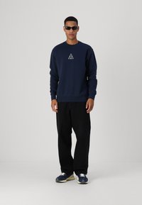 Sudadera marina con cuello redondo y un logo en forma de triángulo blanco. Combinada con pantalones de pierna ancha negros y zapatillas marinas con cordones blancos.