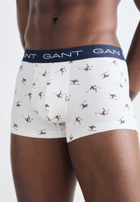 Hvite boxerunderbukser med et skimotiv som viser små figurer i rødt og grønt. Marineblå midjebånd med "GANT"-logo i hvitt. Myk, elastisk stoff.