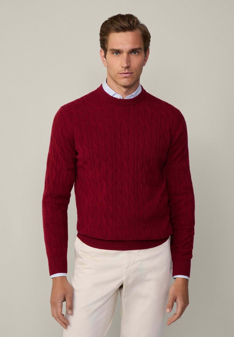 Roter, kabelstrickpullover mit Rundhalsausschnitt, gerippten Bündchen und Saum. Über einem hellblauen Hemd mit Kragen getragen, kombiniert mit beigen Hosen.