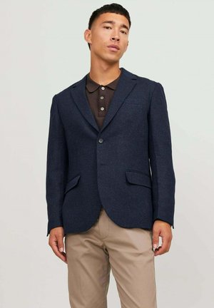 Blazer blu navy in tessuto testurizzato con chiusura a due bottoni, rever a dente e due tasche frontali, abbinate a una polo marrone.