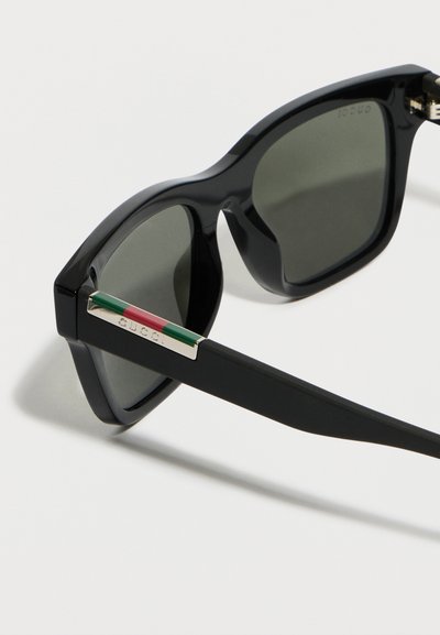 Μαύρα ορθογώνια γυαλιά ηλίου με σκουρόχρωμους φακούς και μαύρο πλαίσιο που διαθέτει πράσινη και κόκκινη μεταλλική ρίγα στα πλαϊνά.