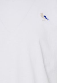 Tricou alb din bumbac, cu un mic păun brodat în bej și albastru în zona stângă a pieptului; design cu decolteu în V și o textură moale.