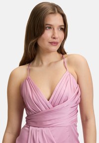 Robe sans manches rose tendre avec un décolleté plongeant, présentant un tissu froncé et un design enroulé mettant en valeur la taille. Texture douce.