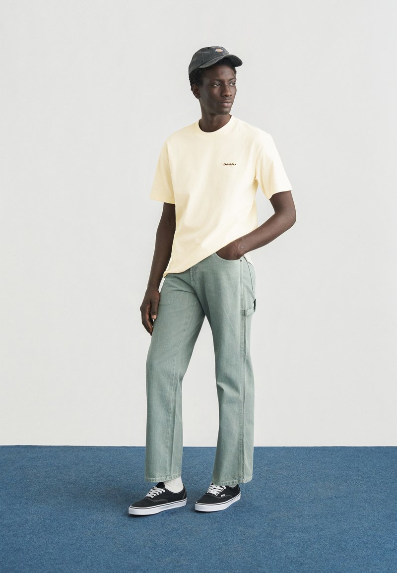 Maglietta a maniche corte giallo chiaro con logo "Dickies" nero, abbinata a pantaloni a gamba dritta verde chiaro e sneakers nere con suola bianca.