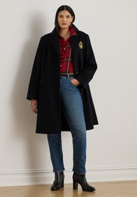 Black wool coat with a structured shape and pointed collar, layered over a red plaid shirt. Worn with blue jeans and black heeled ankle boots.