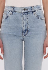 Jeans de mezclilla de lavado claro con cintura alta, bolsillos clásicos, costuras naranjas y un botón plateado en la bragueta.