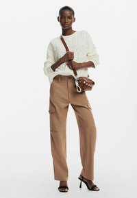 Maglione a trecce crema, pantaloni cargo beige con tasche laterali e sandali neri con tacco. Borsa a tracolla marrone con tracolla e dettagli in metallo.