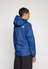 Veste légère bleue avec capuche, fabriquée en matériau résistant à l'eau. Dispose de poignets élastiques et d'un logo dans le dos.
