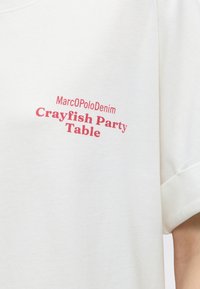 Λευκό ύφασμα με κόκκινο κείμενο "MarcOPoloDenim Crayfish Party Table" τυπωμένο στην περιοχή του στήθους μιας κοντομάνικης μπλούζας.